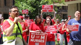 A group of people participate in an outdoor rally in support of University of Michigan nurses. In the foreground, a man in a neon safety vest speaks into a megaphone. Behind him, several individuals in red shirts hold up red and white signs that read "SAFETY STAFFING SOLIDARITY" and "WE SUPPORT U OF M NURSES." To the right, a man in a blue Michigan Medicine t-shirt holds a similar sign. The group is standing in a paved area with green trees and university buildings in the background.