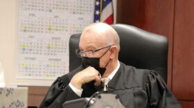 Steuben County Judge Patrick McAllister, wearing his black robes and face mask, sits at his bench with a wall calendar behind him.