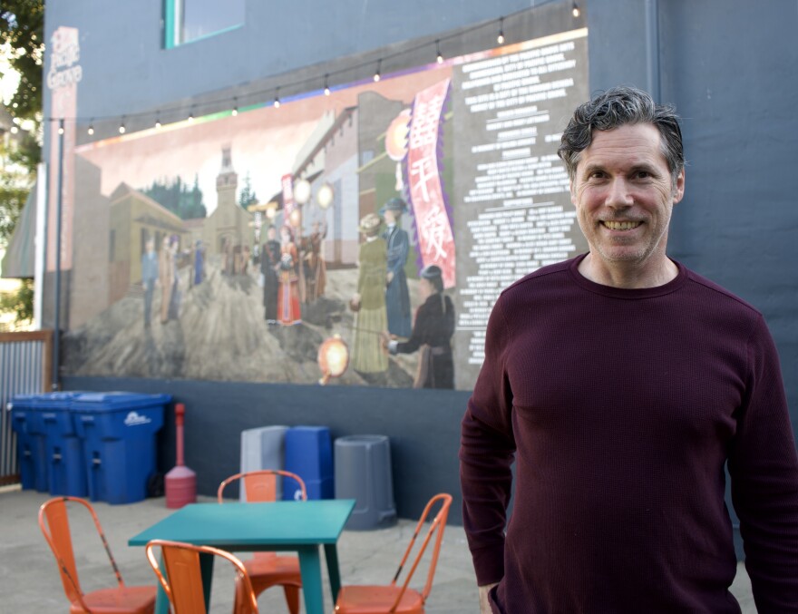 A man stands in front of a mural and table with 4 chairs in an open-air patio. 