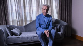 a silver-haired man in a blue suit and white shoes sits on a blue-gray couch