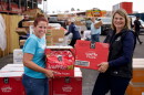Two women, one with red hair in a bun on the left and First Lady Jennie Gordon on the right, smile while holding boxes of waffle fries. In the background, other people move boxes of food at a mobile food pantry.