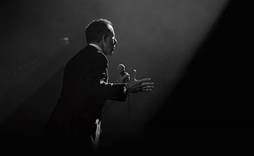 A man wearing a suit faces an unseen audience in a darkened theater while standing under a spotlight and talking into a microphone.