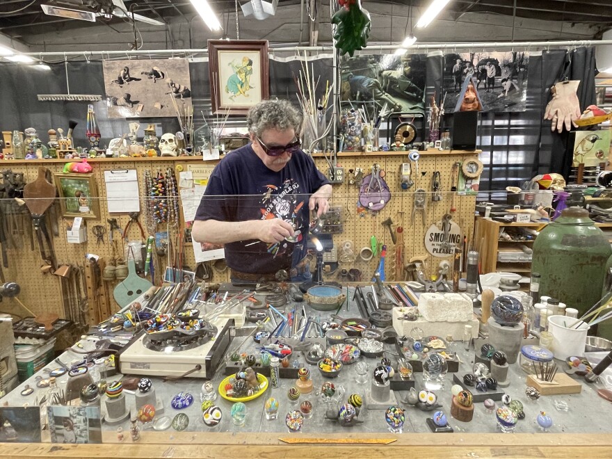 A man wearing safety glasses works a glass marble over flame at a workbench filled with marble making supplies and examples of different styles of marbles. 