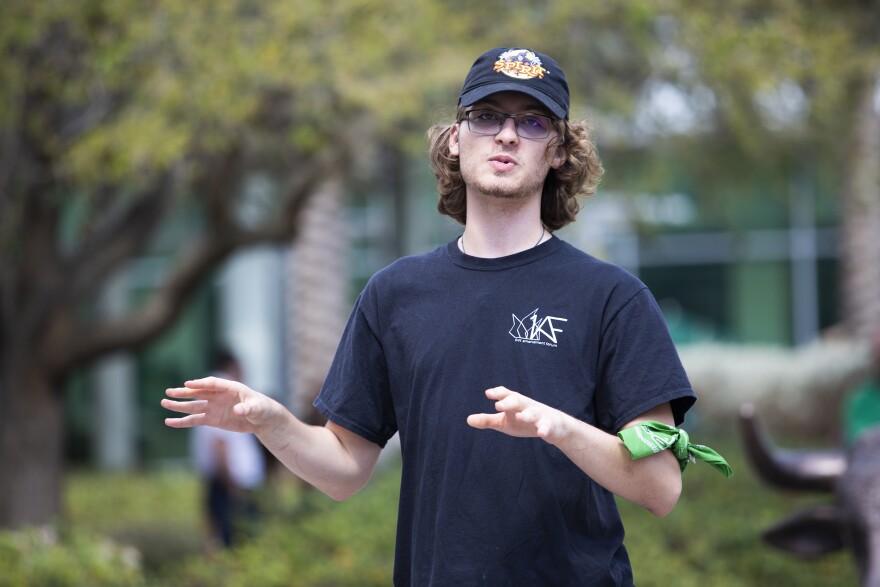 A man wearing a dark blue trucker hat and a green armband speaks to a crowd. 