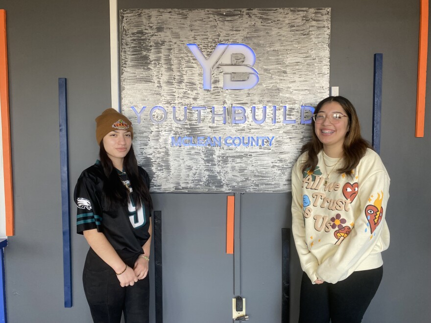 Brittany Ardon stands to the left of a YouthBuild McLean County sign in the lobby of the organization's building. Kristal F stands to the right. Both smile at the camera.