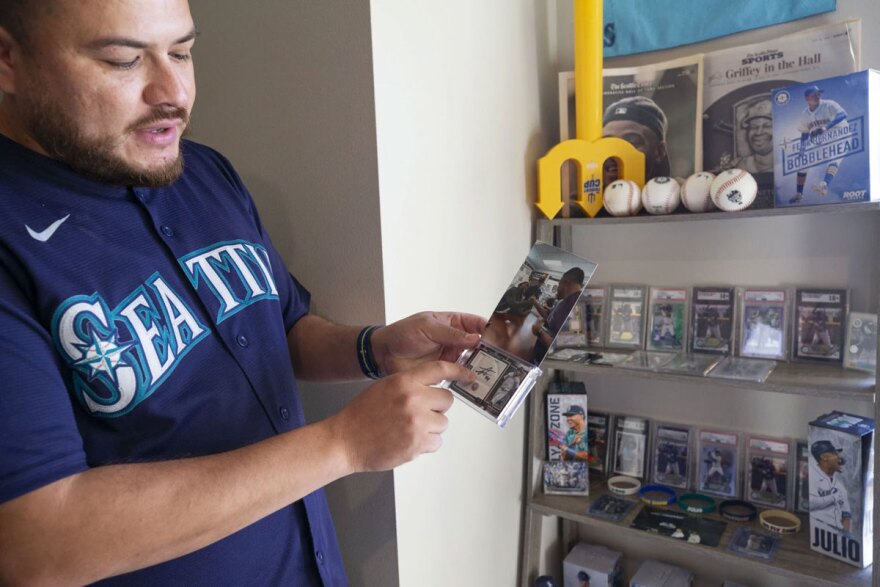 Juan Sanchez points to a signature of Seattle Mariner center fielder Julio Rodriguez on a piece of base in a trading card in his apartment. He is also holding a photo of him meeting Rodriguez during a signing.