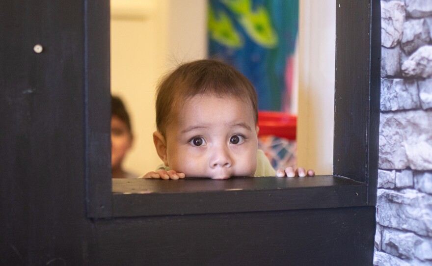 11-month-old Adriel Núñez chews on a window frame. The play area includes multiple sponsored play buildings.
