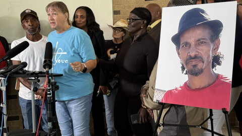 A woman in a blue shirt stands at a microphone next to a photo of a man in a red shirt wearing a hat in front of a group of people
