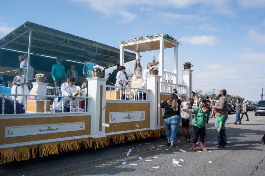 A king and queen stand on an elaborately decorated white and gold float as people shoot fake money at them at a children's parade.