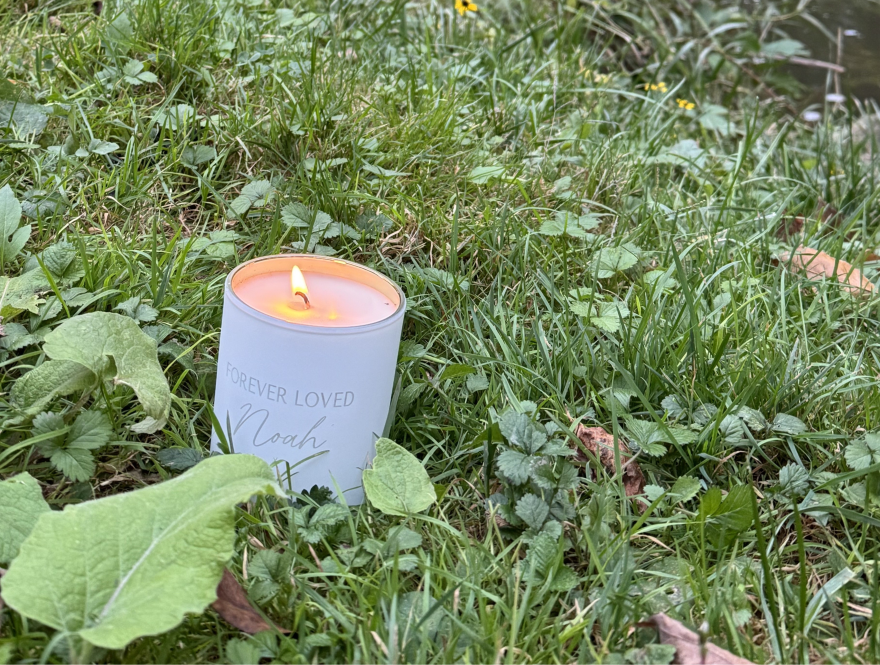 a lit candle that says "forever loved Noah" sits in grass in front of a creek