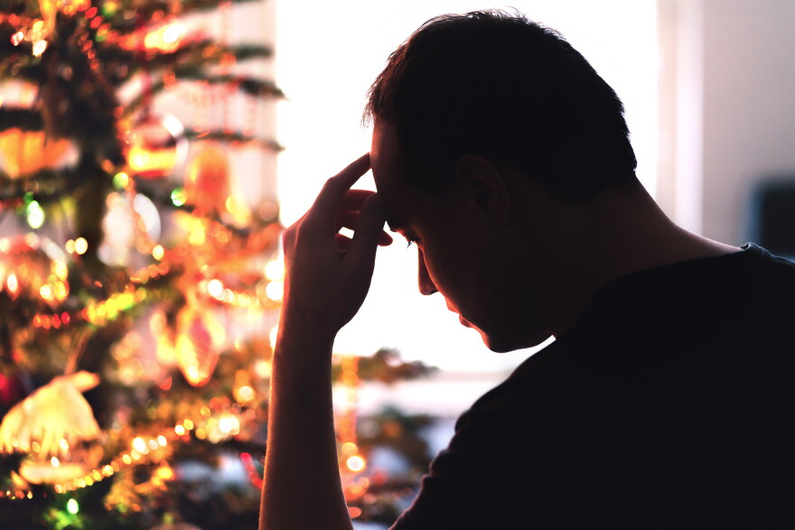 Silhouetted man with his head bowed and fingers pressed to his forehead, standing beside a lit Christmas tree.