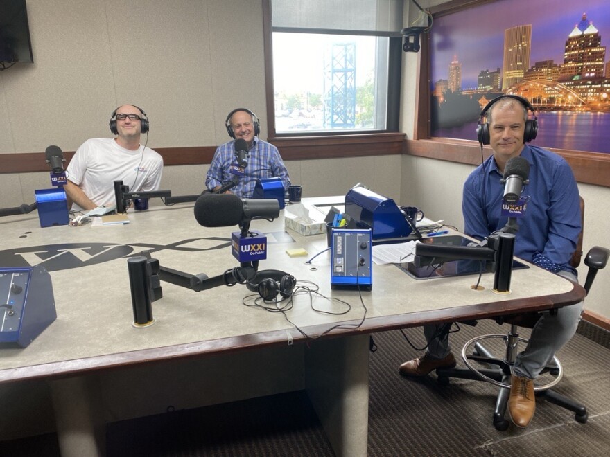 Three men wearing headphones sit at a table in a radio talk studio: the man on the left is bald and is wearing glasses and a white t-shirt; the man in the center is wearing a blue and white plaid long sleeved button down shirt; the man on the right has short dark hair and is wearing a blue long sleeved button down shirt and jeans.