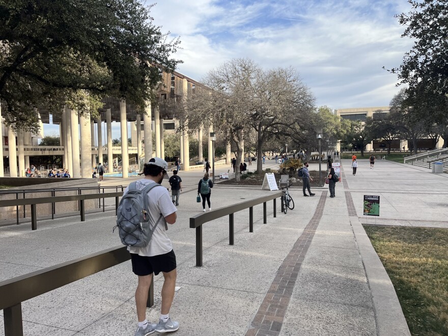 A student in a ball cap and backpack walks down a ramp towards the sombrilla on the main campus of UT San Antonio in March 2026.