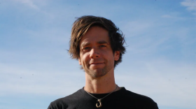 A man with medium-length brown hair stands outside. He wears a black shirt and a hook necklace. He looks at the camera and smiles. 