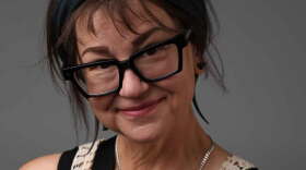 A woman with short brown hair and chunky black glasses smiles at the camera in front of a grey background