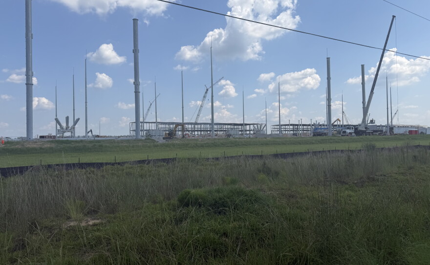 About a dozen large, silver poles are the main focus of this photo. There is grass at the forefront and two steel buildings in the far back distance. 