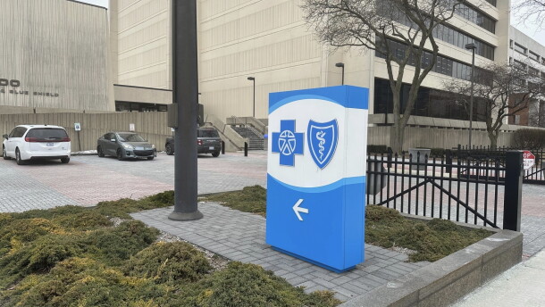 A blue and white sign with the Blue Cross Blue Shield logo stands in a grassy patch in front of a large grey building.