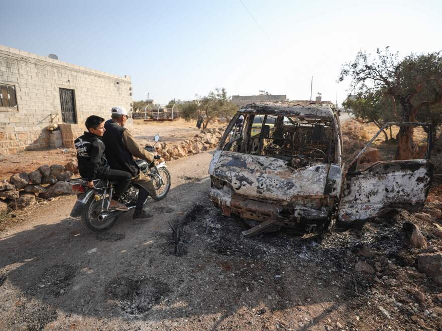 Syrians ride a motorcycle past a burned vehicle near the village of Barisha in the Idlib province of Syria, near where U.S. forces raided the compound of ISIS leader Abu Bakr al-Baghdadi. Local residents tell NPR that noncombatant civilians were killed and injured in the van the night of the U.S. raid.