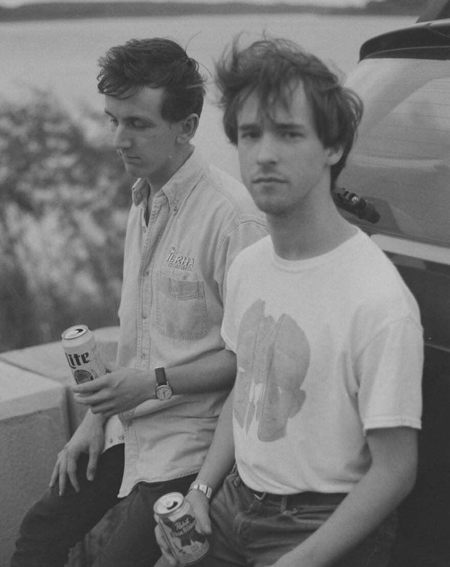 Two men side by side leaning against a car.