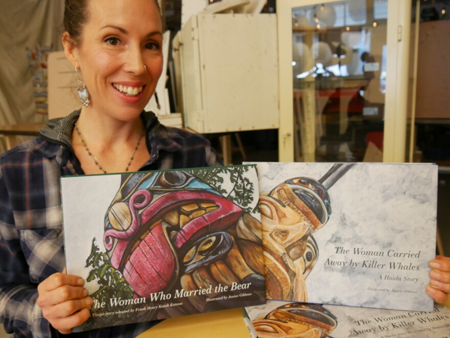 A smiling Haida female artist, Janine Gibbons, holds two books that she worked on for the Baby Raven Reads Program. Both book covers depict artistic renditions of totem pole carvings. (Photo by Joe Viechnicki/KFSK))