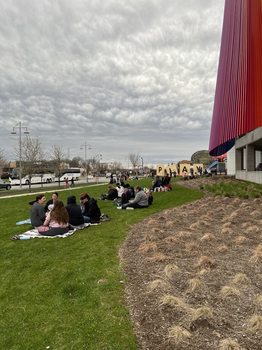 Shortly before totality, families and groups of people await the eclipse on the grass outside of The Strong Museum of Play.