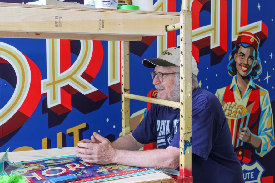A smiling man leans onto a painting scaffold positioned in front of a colorful mural.