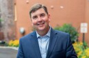 Rep. John Mannion wearing a blue suit jacket and light blue shirt, smiling in front of a brick building.