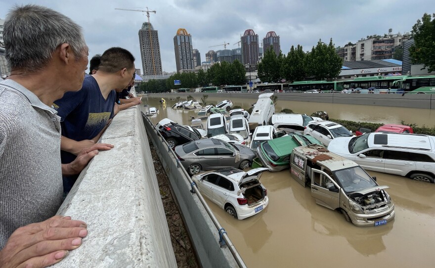 Men peer over a concrete ledge at a pile of cars submerged in muddy floodwaters, with high-rise buildings in the distance.