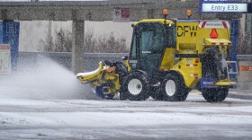 A yellow road machine spits out ice and snow accumulation from the roadway in front of a terminal at DFW Airport. 