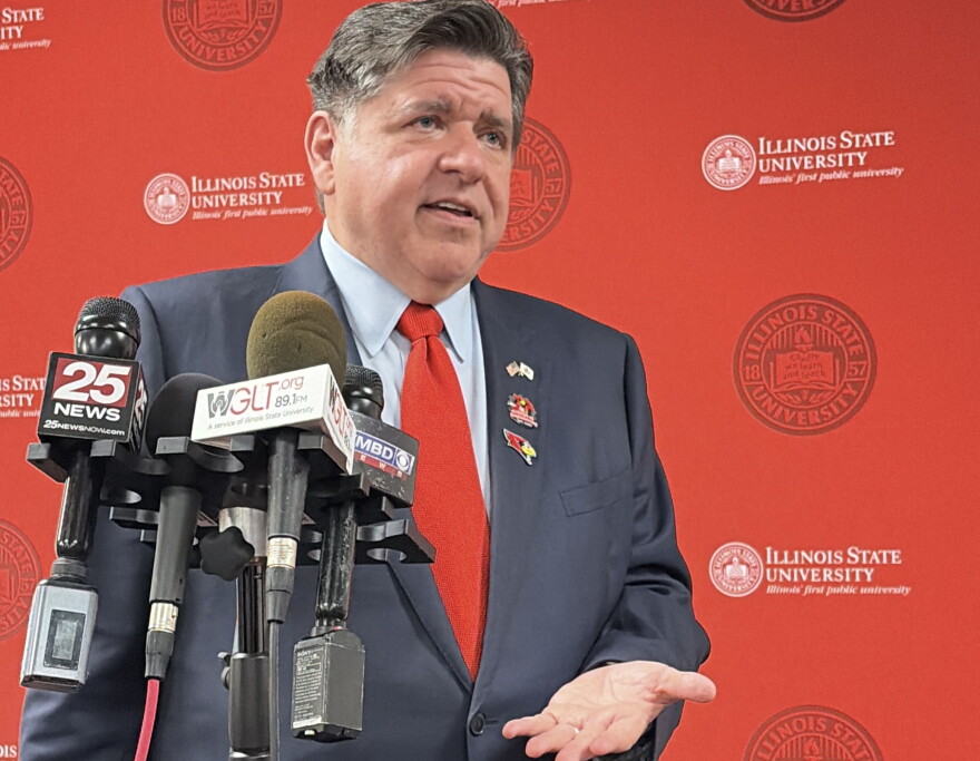 Gov. JB Pritzker speaks into several media microphones standing in front of a red Illinois State University backdrop.