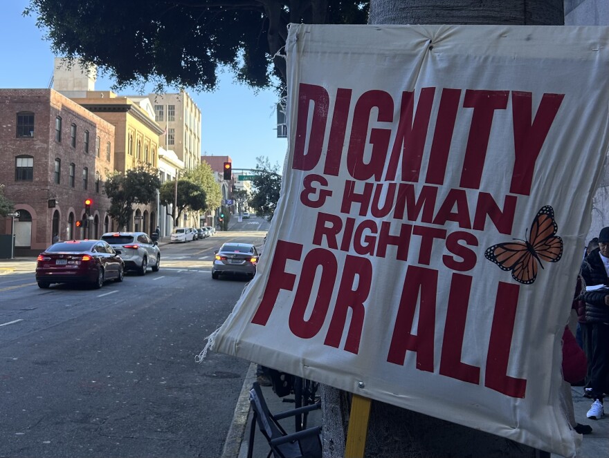 A sign reading "dignity and human rights for all" leans on a tree outside of 630 Sansome Street. 
