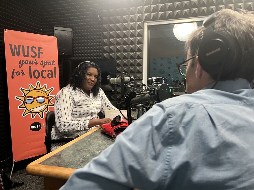 African American woman sits with headphones on near microphone in front of WUSF Your Spot for Local sign. A man's back with headphones over his ears is also seen sitting across from her next to another microphone.