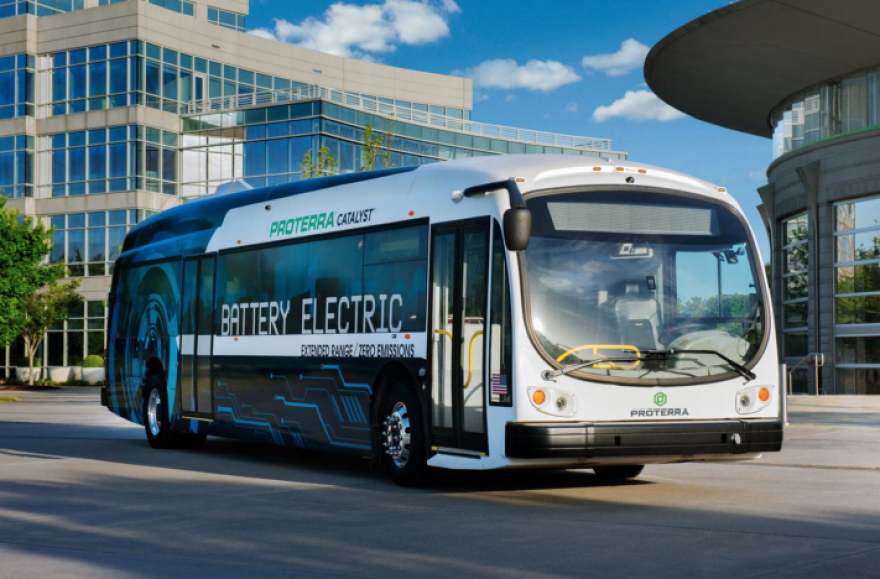 A Proterra battery-electric bus is parked in an urban environment, with modern glass buildings in the background. The bus is white with the words "Battery Electric" prominently displayed on its side.