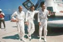 Three men in white jumpsuits stand in front of a spacecraft. They appear to be talking with someone out of frame to their left.