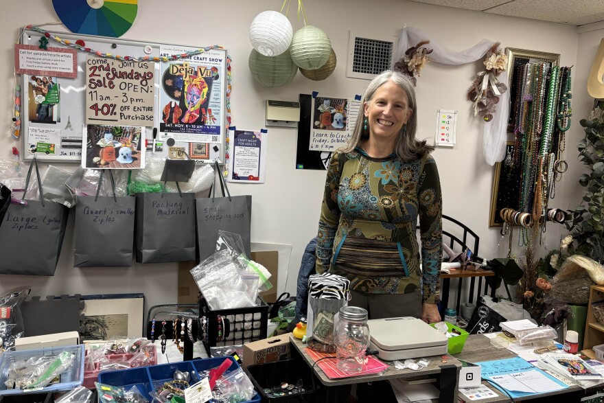 Sue McCracken standing behind a desk filled with bins and jars full of small objects, a scale, a card reader. Behind her are posters and bags labeled as holding different sized bags. strands of beads hang behind her. 