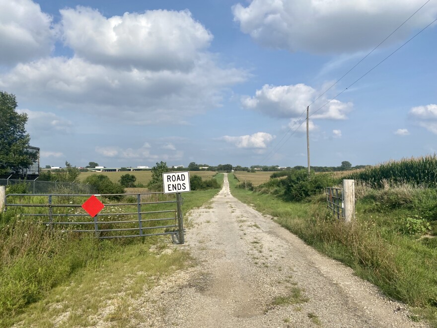 Looking through an open metal gate with a red diamond and a "road ends" sign posted, gravel road dead-ends in the distance. Beyond that, a busy expressway is barely seen in the distance.