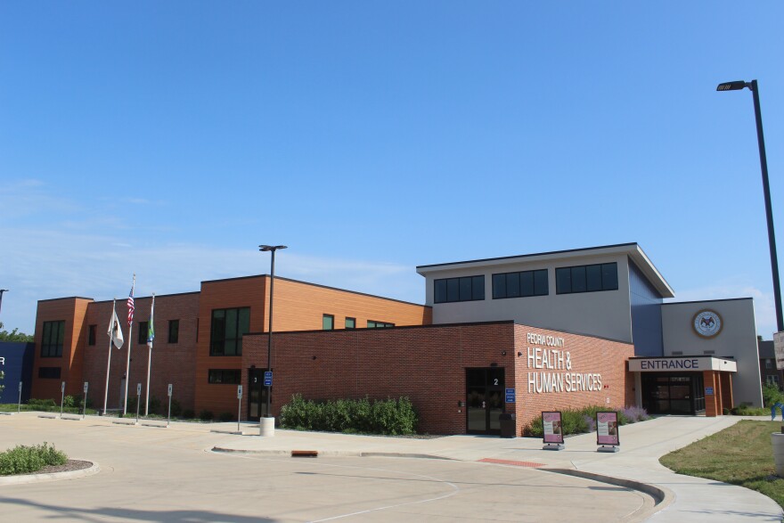 Exterior photo of the Peoria County Health & Human Services building, a modern two-story structure combining red brick, warm wood-toned paneling, and gray metal siding with large black-framed windows. Signage reads "Peoria County Health & Human Services" above a brick facade, with a covered entrance, the county's circular seal on the building's tower section, and an American flag, state flag, and county flags on poles out front. WIC program signs stand near the entrance under a clear blue sky.