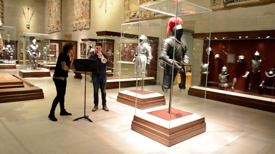 Woman stands next to man playing trumpet in museum armor court
