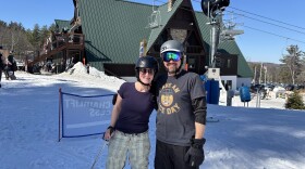 Two skiers in t-shirts pose for a photo in front of a ski lodge. 