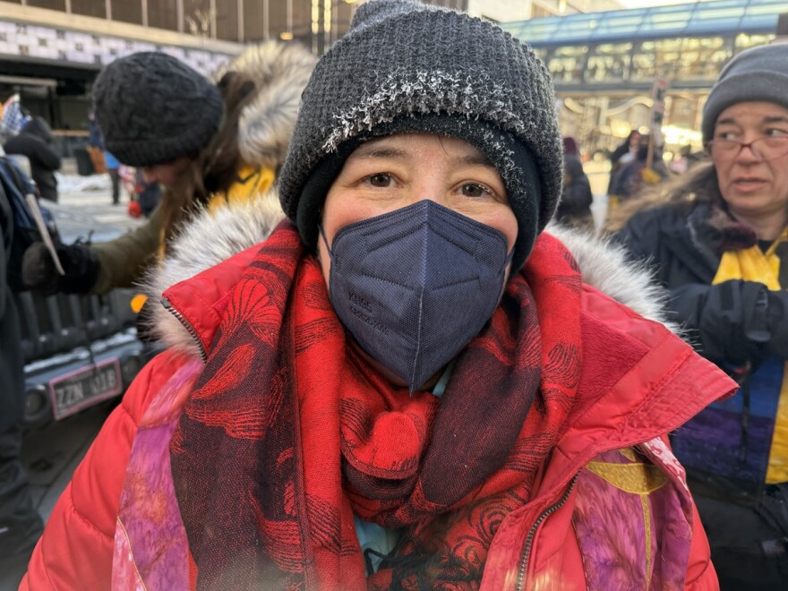 Woman in a red coat and wearing a wool hat and facemask and standing in front of a group of people on a city street