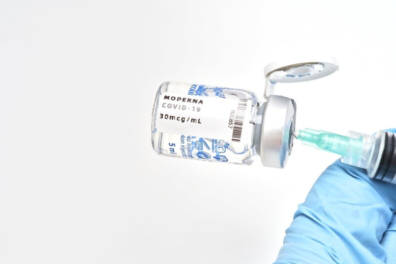 A large vial of the Moderna COVID-19 vaccine with a syringe sticking out the top hangs sideways in front of a white background.