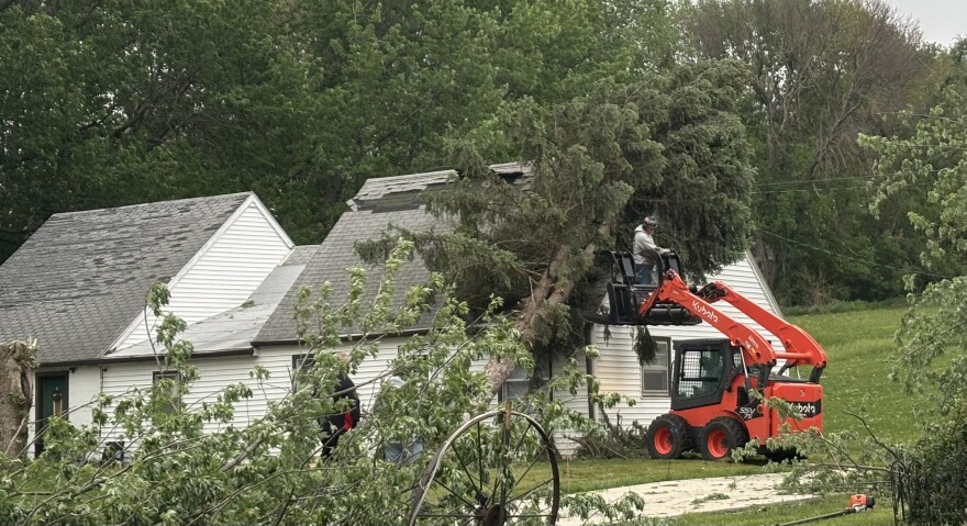 A worker uses a skid loader to remove a large diameter tree from a home in Moville, Iowa, on May 18, 2026, following heavy area storms. (Bret Hayworth, Siouxland Public Media News)
