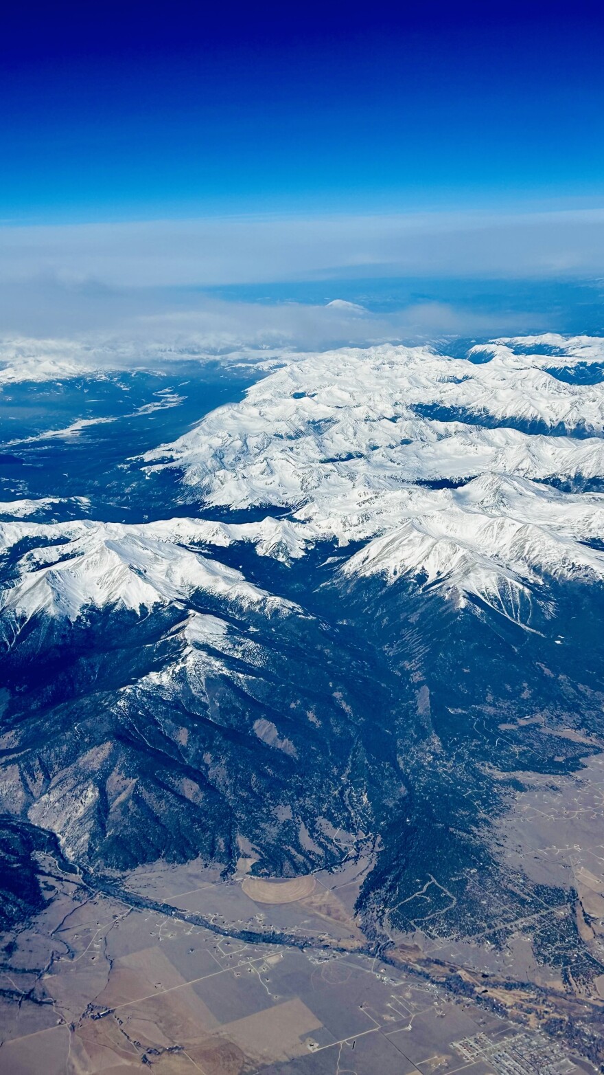 Snow covers the very tops of large mountain peaks in an aerial photograph. The cities at the base of the mountains are dry and surrounded by brown fields. 