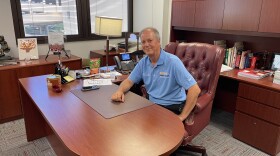 A man in a blue polo sits at his desk and smiles at the camera