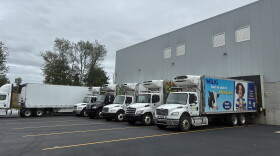 Outside of a warehouse with six food distribution trucks parked outside of it 