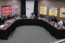 A group of people sit around a large U-shaped table in a meeting room with bulletin boards on the walls and “Peoria Public Schools” on a banner behind the center person.