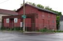 A brick building, painted red, with painted red boards over the windows behind a chain-link fence. In front of it is a light pole with street signs for Ridge Road and Gravel Road attached.