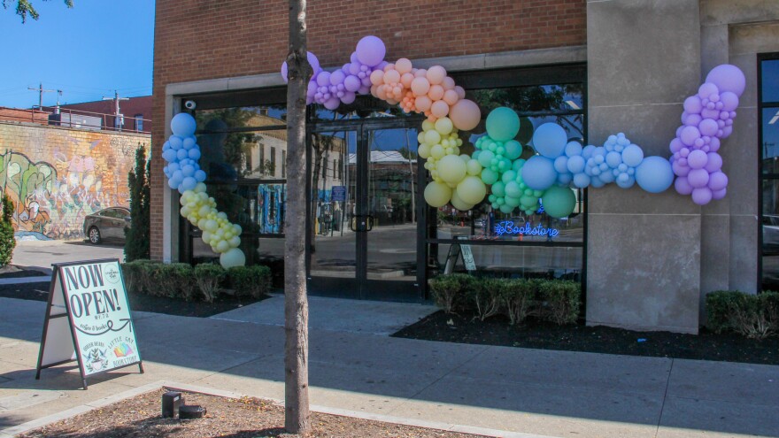 The outside of a storefront is covered rainbow balloons. A sidewalk sign reads "Now open!"