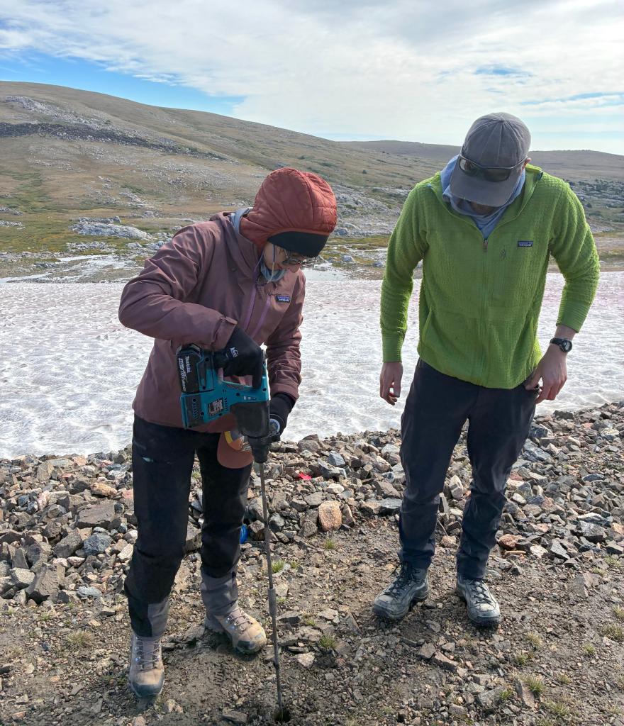 Two people in cold-weather gear stand on rocky ground near an ice patch. One person is drilling into the ground with a very long drillhead.