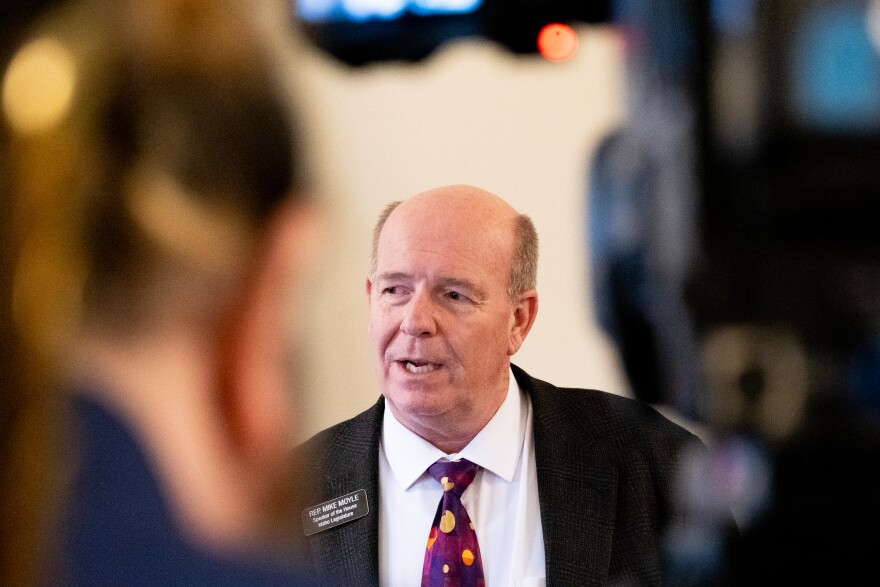 A balding man in a gray suit framed between a television camera and the camera operator.